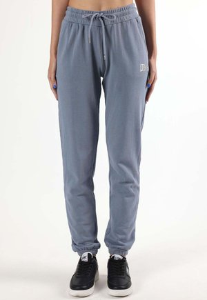 Femme portant un pantalon de jogging gris avec taille et poignets élastiques, debout avec les mains détendues le long du corps, associée à des baskets noires.