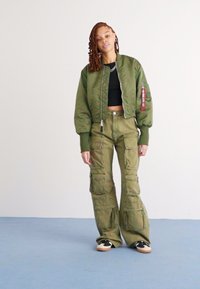 Groene bomberjack met een cropped ontwerp, gecombineerd met olijfgroene cargobroek met meerdere zakken. Een zwart top en zwarte sneakers maken de outfit compleet.