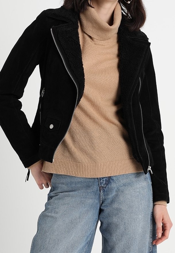 Giubbotto in ecopelle nera da motociclista con colletto in shearling, zip argentate e tasche laterali, indossato su un dolcevita beige e jeans blu.