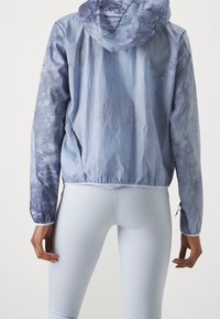 Lys blå vindjakke med tie-dye mønster, lynlåslommer og elastiske manchetter, iført over lys blå leggings.