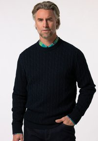 Granatowy sweter z splotem w warkocz, z prążkowanymi mankietami i dołem. Noszony na zielonej i niebieskiej koszuli w kratę z kołnierzykiem, o gładkiej teksturze.