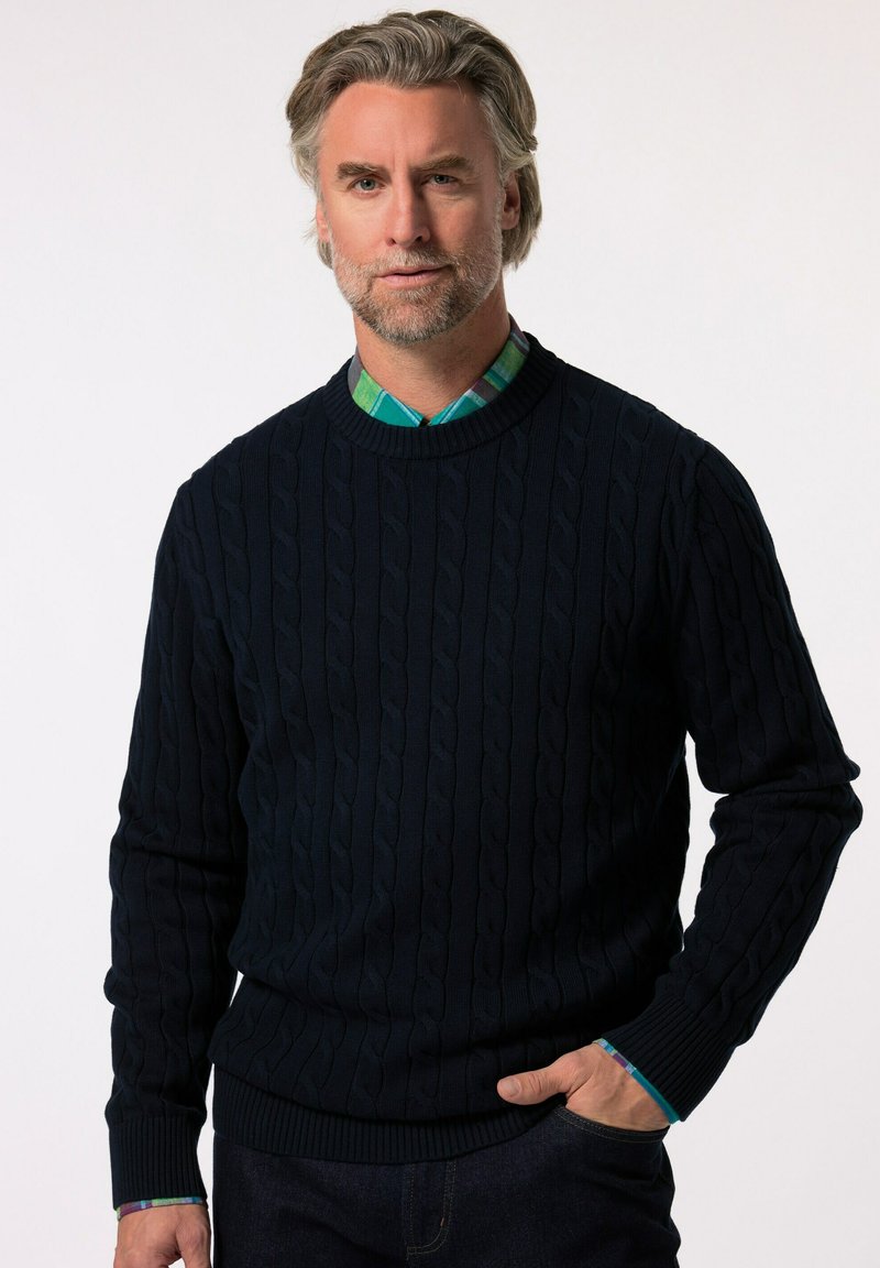 Granatowy sweter z splotem w warkocz, z prążkowanymi mankietami i dołem. Noszony na zielonej i niebieskiej koszuli w kratę z kołnierzykiem, o gładkiej teksturze.