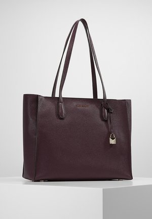 Tummanvioletti nahkainen olkalaukku, jossa on kaksinkertaiset olkahihnat, kullanvärinen logo ja roikkuva pieni lukko, valkoisella jalustalla.