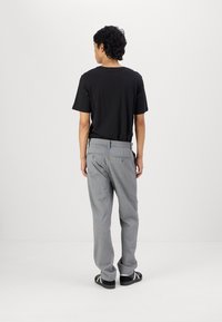 Schwarzes T-Shirt, graue Hose, elastischer Bund mit Gesäßtaschen. Die Hose hat einen geraden Schnitt und eine dezente Textur. Einfache Sandalen runden den Look ab.