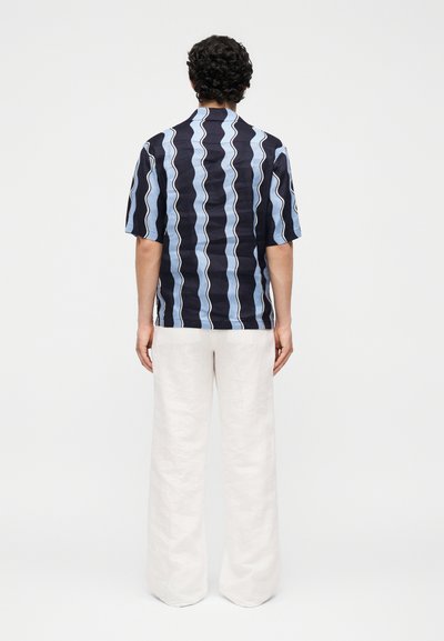 Άνδρας με σγουρά μαλλιά που φοράει μπλε σκούρο και φωτεινό μπλε ριγέ πουκάμισο με κοντά μανίκια και λευκό παντελόνι με φαρδιά μπατζάκια, στέκεται με την πλάτη στραμμένη.