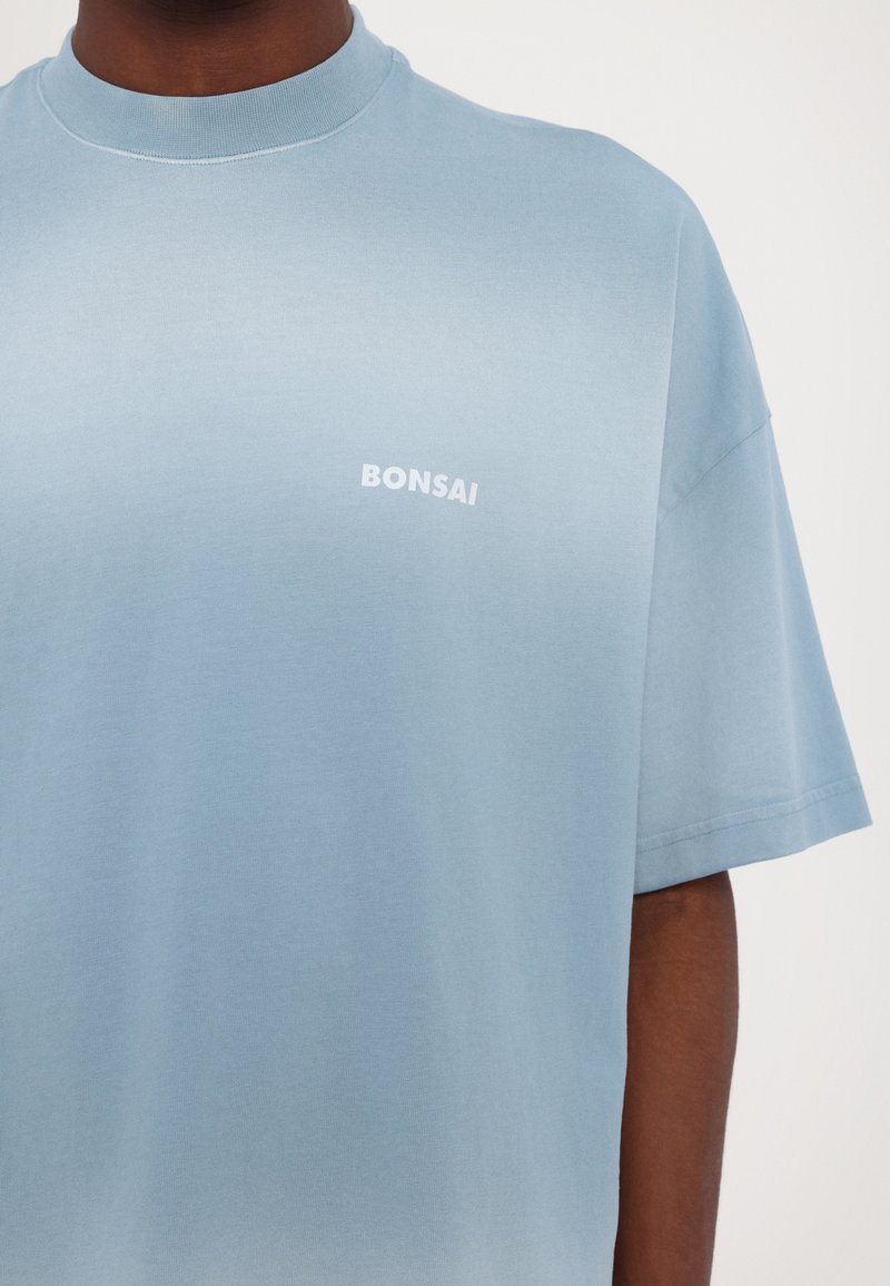 Himmelblaues Baumwoll-T-Shirt mit kurzen Ärmeln, das über einen runden Halsausschnitt verfügt und auf der linken Brustseite weißen Text "BONSAI" zeigt.