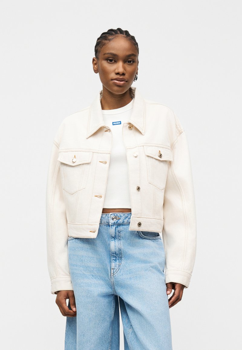 Νεαρή γυναίκα με πλεγμένα μαλλιά, φορώντας ένα κρεπ κρεμ μπουφάν πάνω από ένα λευκό κρεπ τοπ και ψηλόμεσα ανοιχτό μπλε τζιν.