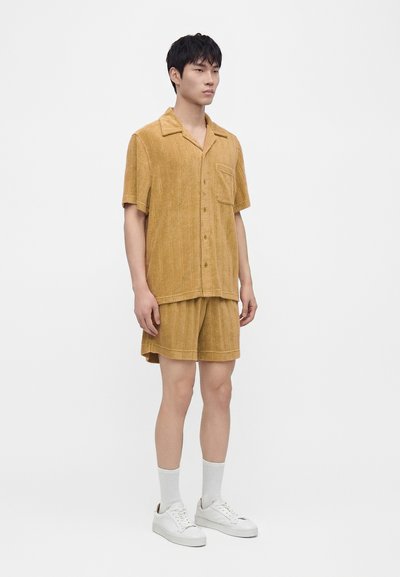 Homme debout portant une chemise moutarde à manches courtes boutonnée et un short assorti avec des chaussettes blanches et des baskets blanches sur fond uni.
