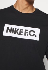 Sort bomulds T-shirt med et hvidt rektangulært grafik, hvor der står "NIKE F.C." med fede sorte bogstaver, rund hals og korte ærmer.
