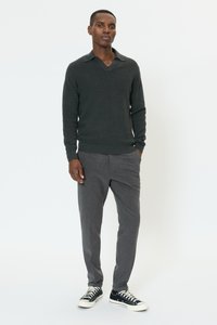 Mørkegrøn strikket sweater med V-hals, kombineret med grå skræddersyede bukser og sorte sneakers med hvide detaljer, stående mod en hvid baggrund.