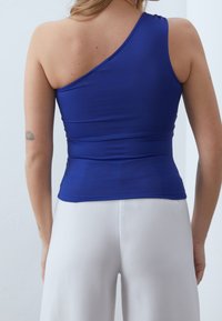 Asymmetrisk blå topp gjord av elastiskt tyg, med en tight passform och drapering på ena axeln, tillsammans med vita byxor.