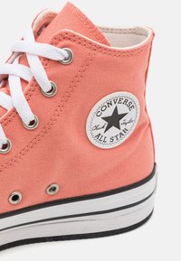 Zapatillas Converse de caña alta en lona coral con cordones blancos, ojales plateados y un parche de estrella en blanco y negro en el costado.
