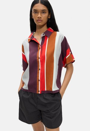 Kobieta ubrana w koszulę zapinaną na guziki z pionowymi paskami w kolorze bordowym, czerwonym, pomarańczowym i białym, połączona z luźnymi czarnymi szortami, stojąca z rękami w kieszeniach.