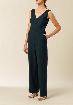 Donkergroen jumpsuit met een diepe V-hals en wijde pijpen, voorzien van een getailleerde taille en zijzakken. Gecombineerd met lichtgekleurde chunky sandalen.