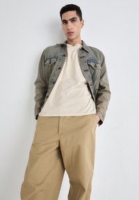 Jeansjacke in Hellblau, cremiges T-Shirt und beigefarbene Hose. Die Jacke verfügt über klassische Taschen und hat einen leicht verblassten Look.