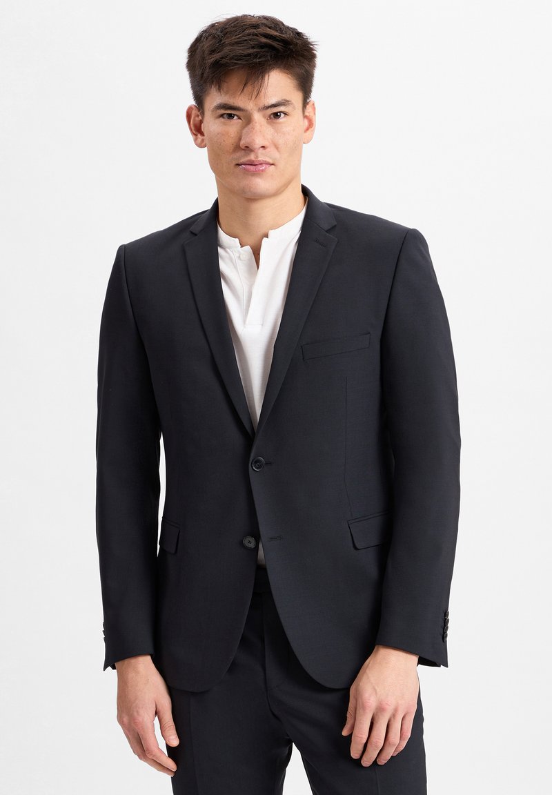 Costume sombre bleu marine pour homme avec une veste à un bouton, coupe ajustée, revers crantés et une poche poitrine. Porté par-dessus une chemise henley blanche.