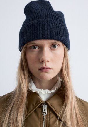 Navy ribbed knit beanie in wool, fitted with a unique peak design. Worn with a brown bomber jacket and a white ruffled collar underneath.