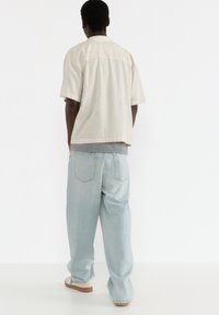Hellbeige Hemd mit kurzen Ärmeln und geradem Schnitt, kombiniert mit lockeren hellblauen Jeans. Das Outfit weist ein geschichtetes Design und weiche Texturen auf.