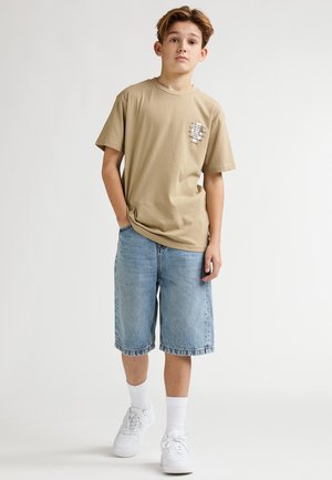 T-shirt di cotone beige con una piccola stampa di testo, abbinata a pantaloncini di jeans azzurri, sneakers bianche e calzini bianchi alla caviglia. Design semplice.