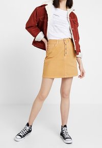 Chaqueta de mezclilla con forro de sherpa, camiseta gráfica blanca y falda de mezclilla color mostaza con botones en el frente. Combinada con zapatillas negras de caña alta.