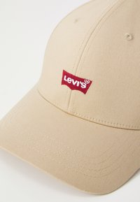 Beige pet gemaakt van gestructureerde stof met een rood geborduurd Levi's logo, een gebogen rand en zichtbare stikselaccenten.