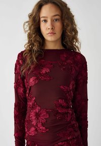 Burgunderfarbener Langarm-Top mit floraler Stickerei, ausgestattet mit einer transparenten Überlagerung und strukturiertem Stoff. Mit erhabenen Blumenmustern verziert.