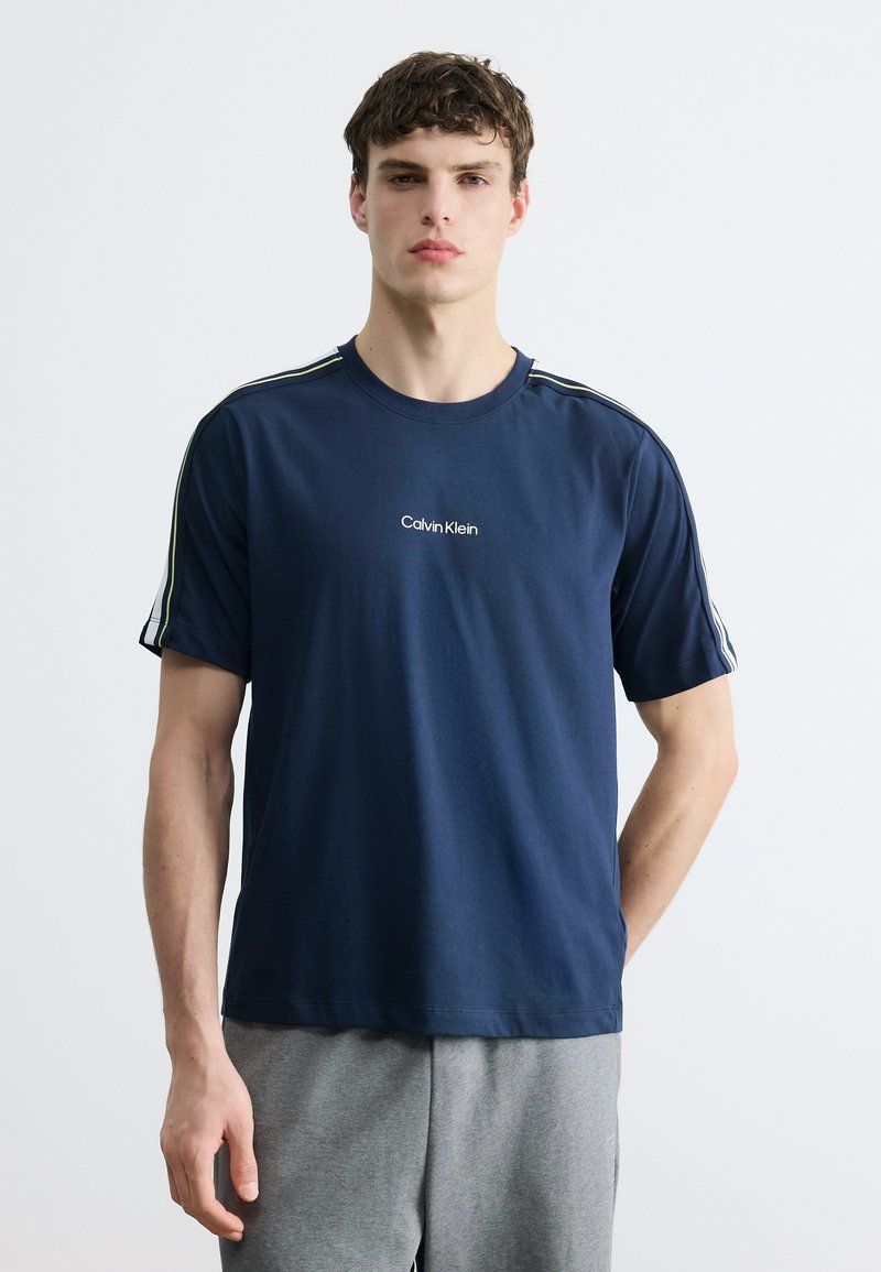 Νεαρός άνδρας που φοράει μπλε σκούρο t-shirt Calvin Klein με λευκές ρίγες στα μανίκια και γκρι παντελόνι, στέκεται μπροστά σε λευκό απλό φόντο.