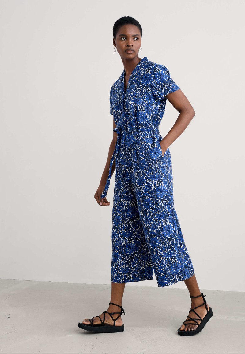 Zils ziedu raksts kopējais apģērbs, kas izgatavots no viegla auduma; ar īsām piedurknēm, jostu viduklī un platām apgrieztām kājām. Melnas siksniņas sandales.