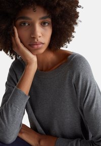 Szary, dziany sweter z luźnym dekoltem i prążkowanymi mankietami, noszony przez osobę o kręconych włosach, która opiera podbródek na jednej ręce.