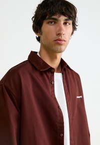 Μαρούλι πουκάμισο με κολάρο, φτιαγμένο από υφασμάτινο υλικό, με λευκό λογότυπο στο στήθος, φορεμένο πάνω από λευκό T-shirt με στρογγυλή λαιμόκοψη.