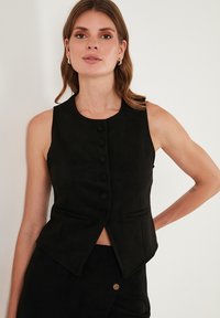 Vesta neagră fără mâneci cu decolteu rotund, închidere cu nasturi și buzunare în față. Fabricată dintr-un material neted, texturat, cu o croială adaptată.