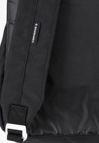 Sac à dos noir avec un tissu texturé, une sangle réglable et une boucle blanche. Comprend une étiquette de marque avec le logo "Converse".