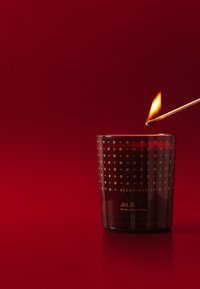 Duftlys i en mørk glasbeholder med gyldne geometriske mønstre, mærket "JUL" af Skandinavisk, med en tændt kamp, der antænder vægen.