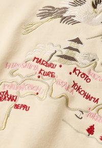 Tessuto beige con una mappa del Giappone ricamata, con luoghi rappresentati in vari colori. Dettagli includono alberi, montagne e una struttura pagoda.