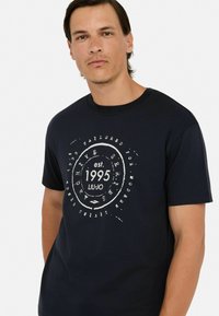 Tamno plava pamučna majica s kratkim rukavima i okruglim izrezom. Sadrži bijeli kružni grafički uzorak s natpisom "osn. 1995 LIUJO" u sredini.