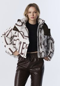 Jeune femme portant une doudoune blanche avec un imprimé abstrait marron et un pantalon en cuir marron foncé, se tenant les mains dans les poches.