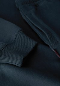 Tejido de sudadera azul oscuro con puños acanalados y mangas largas. Presenta una textura suave y una pequeña etiqueta roja en la costura lateral.