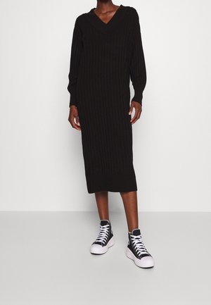 Person iført en sort langærmet, ribstrikket midi-kjole med V-hals og sorte høje sneakers med hvide såler og snørebånd.
