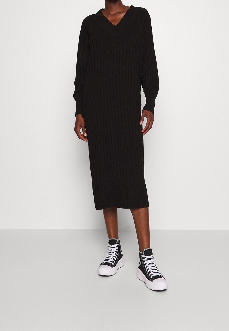 Personne portant une robe midi noire à manches longues en maille côtelée avec un col en V et des baskets montantes noires avec semelles blanches et lacets blancs.