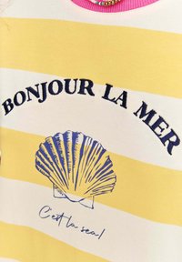 Žluté a bílé pruhované tričko s modrou grafikou mušle. Text zní "BONJOUR LA MER" a "C'est la seal." Má růžový výstřih.