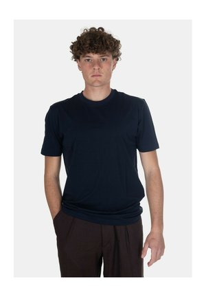 Giovane uomo con capelli ricci che indossa una t-shirt blu scuro semplice e pantaloni scuri, in piedi con una mano in tasca su sfondo bianco.