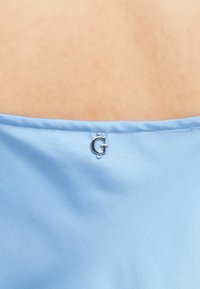 Tecido azul claro com uma textura suave, apresentando um detalhe em forma de logótipo "G" prateado perto da borda superior. Design simples, sem padrões ou acentos adicionais.