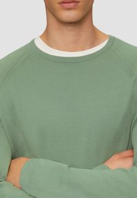 Pull long à manches longues vert clair en matière côtelée, présentant un col rond blanc et des manches raglan avec des détails de couture subtils.