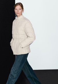 Femme portant une veste matelassée beige clair et un jean bleu marchant devant un fond moitié noir, moitié blanc.