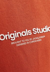 Tejido de algodón rojo con texto blanco que dice "Originals Studio" y detalles sobre la marca y la ubicación del diseño. Textura suave.