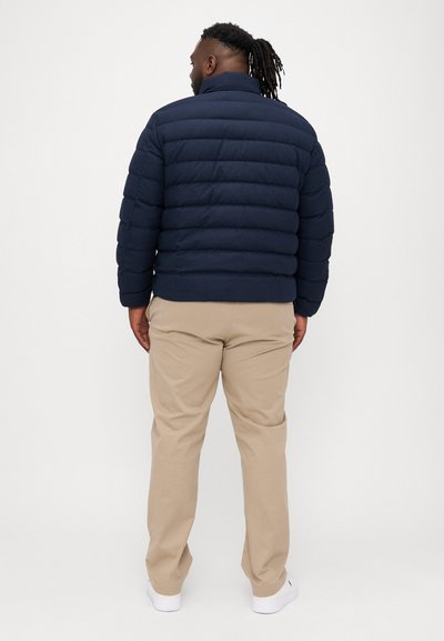 Tamna puffer jakna s horizontalnim šavovima, visokim ovratnikom i kratkim rukavima. Bež hlače i bijele tenisice upotpunjuju outfit.