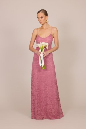 Donna in un abito senza maniche in pizzo malva che tiene un bouquet di calle bianche legate con un nastro bianco su uno sfondo beige.
