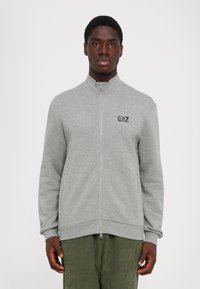 Mann iført en grå zip-up jakke med EA7 Emporio Armani-logo på brystet og olivengrønne teksturerte bukser, står mot en hvit bakgrunn.
