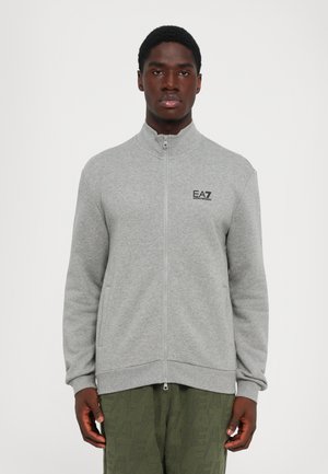Mann iført en grå zip-up jakke med EA7 Emporio Armani-logo på brystet og olivengrønne teksturerte bukser, står mot en hvit bakgrunn.