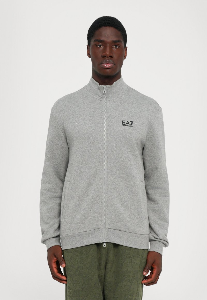 Mann iført en grå zip-up jakke med EA7 Emporio Armani-logo på brystet og olivengrønne teksturerte bukser, står mot en hvit bakgrunn.
